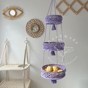 Sweet purple macrame fruit basket with three circular tiers, ornate weaving, and tassels, hung by a wooden ring, showcasing fresh fruits and plants in a cozy home setting with decorative accents