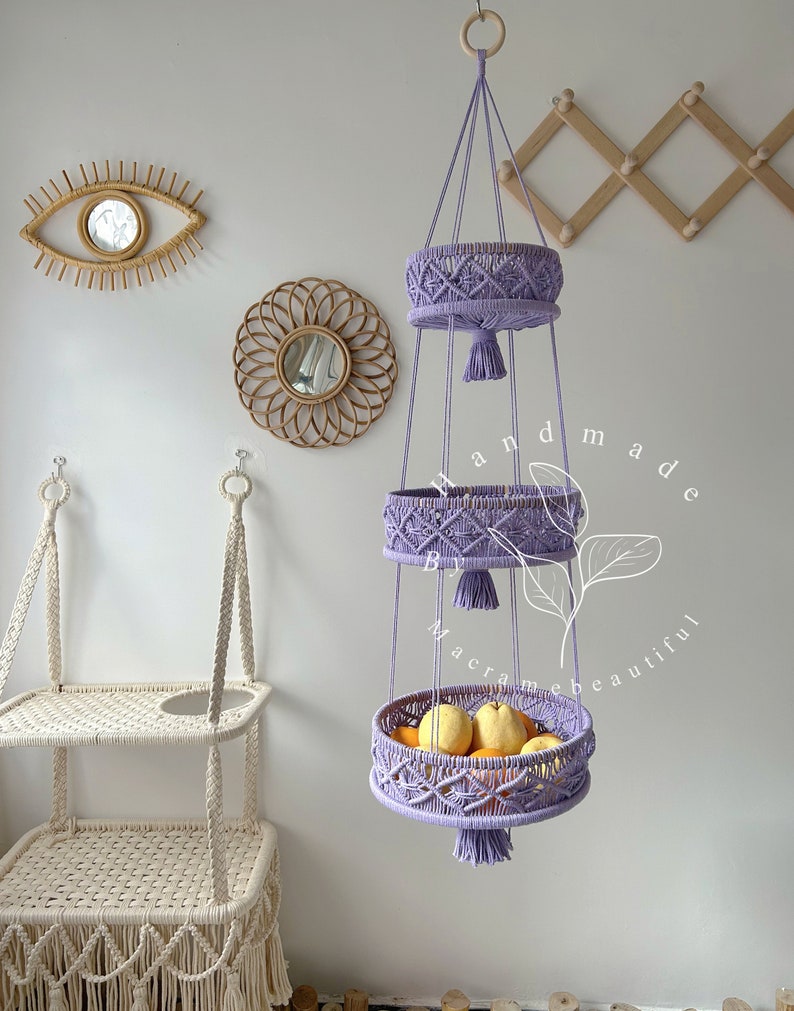 Sweet purple macrame fruit basket with three circular tiers, ornate weaving, and tassels, hung by a wooden ring, showcasing fresh fruits and plants in a cozy home setting with decorative accents