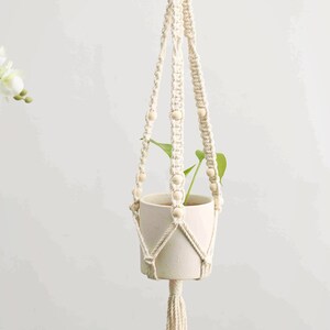 May include: Cream-colored macrame plant hanger with a wooden ring and beads. The woven design holds a white ceramic pot with a green plant. A tassel hangs from the bottom. The letter "B" is visible in the background.