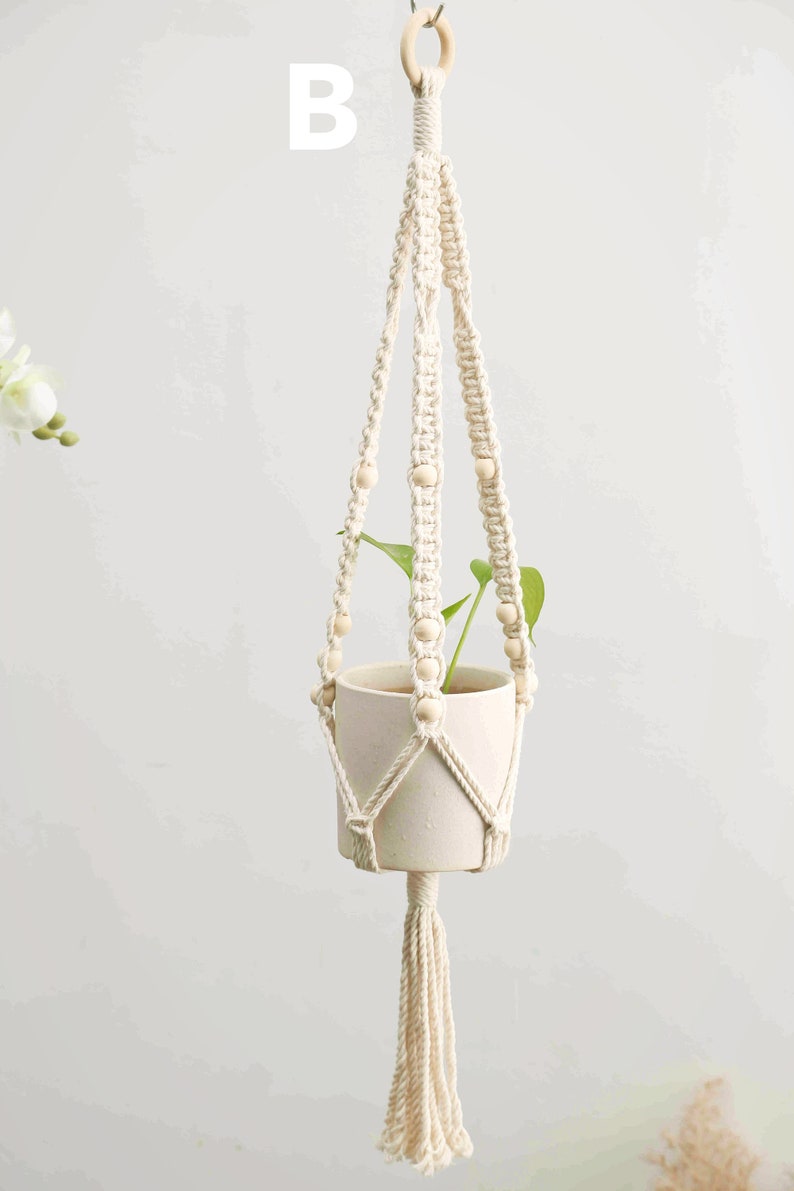 May include: Cream-colored macrame plant hanger with a wooden ring and beads. The woven design holds a white ceramic pot with a green plant. A tassel hangs from the bottom. The letter "B" is visible in the background.