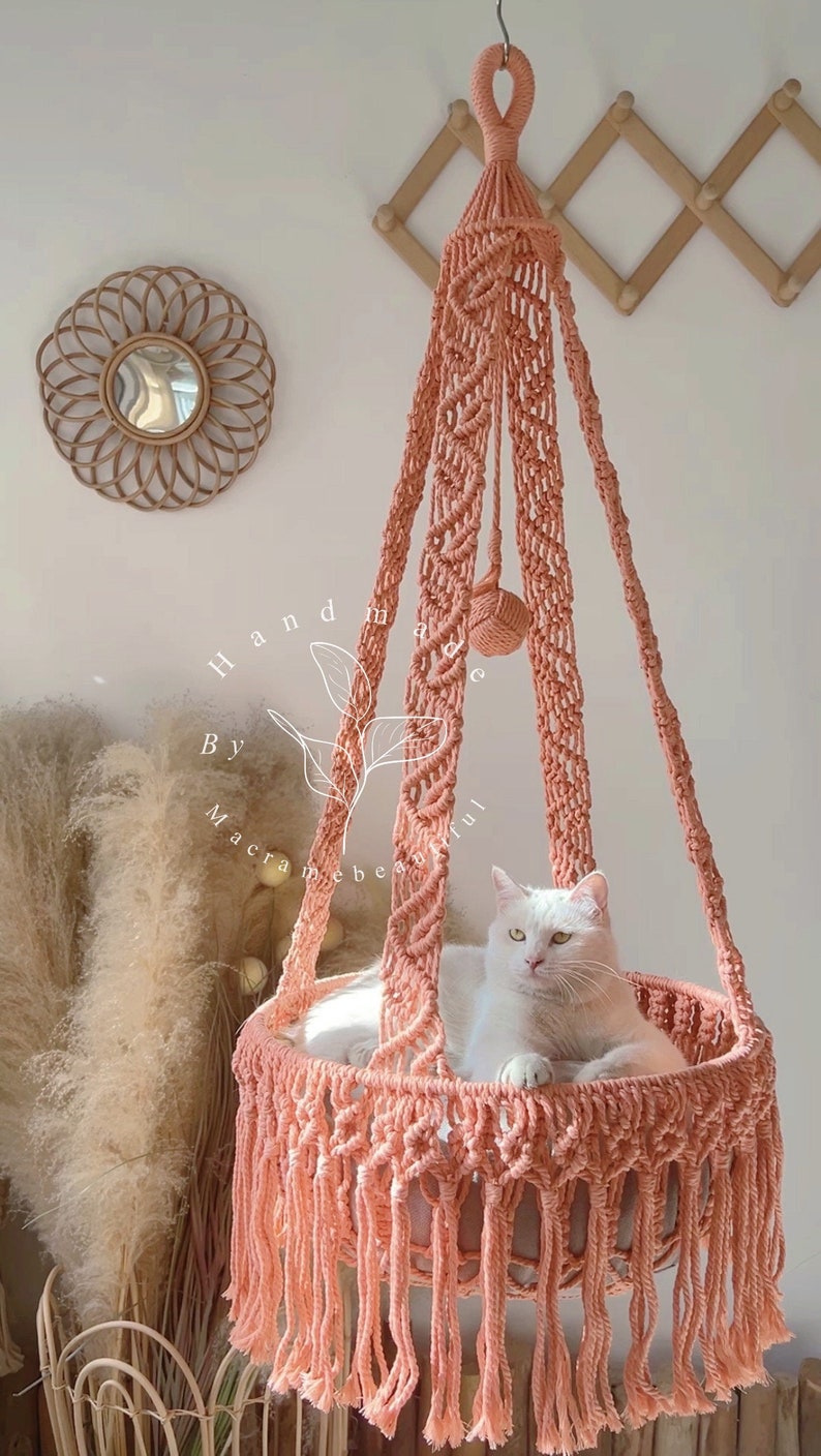 May include: A peach-colored macrame cat bed with a white cat inside. The hanging bed is suspended from a hook and features intricate knotting and fringe. The background includes a decorative mirror and dried plants.