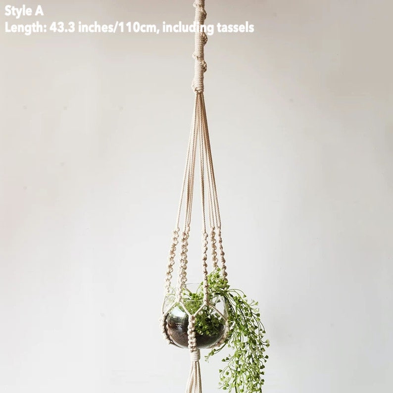 May include: A beige macrame plant hanger with a glass terrarium holding greenery. The hanger is 43.3 inches long, including tassels. The design features knotted rope and decorative beads, creating a bohemian aesthetic. The plant hanger is suspended against a white background.