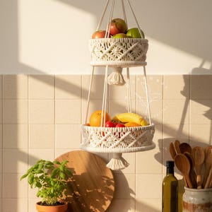 Handmade 2-tier macrame fruit basket hanging in a bright kitchen, woven cotton shelves holding apples, bananas, oranges, and tomatoes, boho farmhouse decor for organized countertop storage and natural kitchen styling