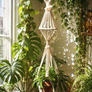Puede incluir: Un colgador de plantas de macramé color crema con un patrón de nudos geométricos en forma de diamante y un lazo superior. El colgador sostiene una maceta de terracota con una planta verde y una esfera de cristal transparente en la parte superior.