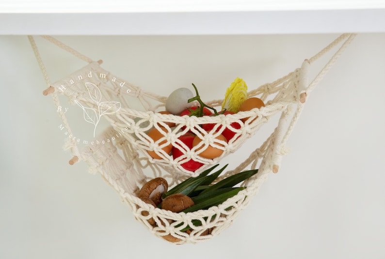 Overhead view of a two-tiered macrame fruit hammock filled with fresh bananas, tomatoes, and apples, showcasing the detailed knotwork, providing a stylish and bohemian kitchen storage solution