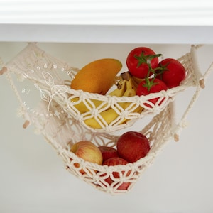 Overhead view of a two-tiered macrame fruit hammock filled with fresh bananas, tomatoes, and apples, showcasing the detailed knotwork, providing a stylish and bohemian kitchen storage solution