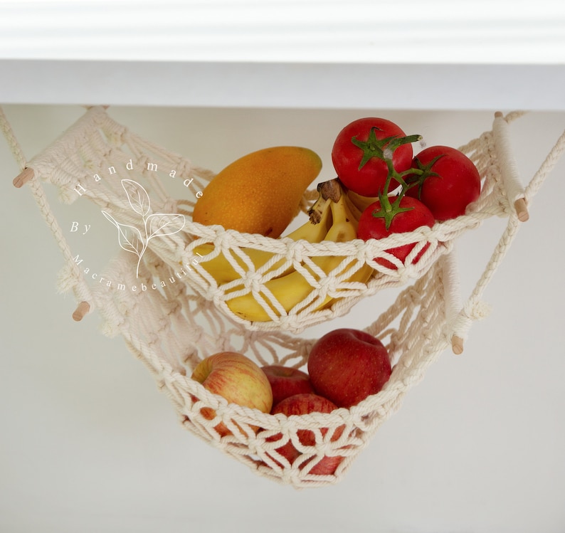 Overhead view of a two-tiered macrame fruit hammock filled with fresh bananas, tomatoes, and apples, showcasing the detailed knotwork, providing a stylish and bohemian kitchen storage solution
