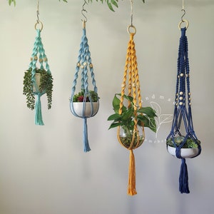 May include: Four macrame plant hangers in shades of teal, blue, yellow, and navy blue, each holding a different plant. The hangers are suspended from wooden rings and feature wooden beads. The plants are in white ceramic pots or glass globes.