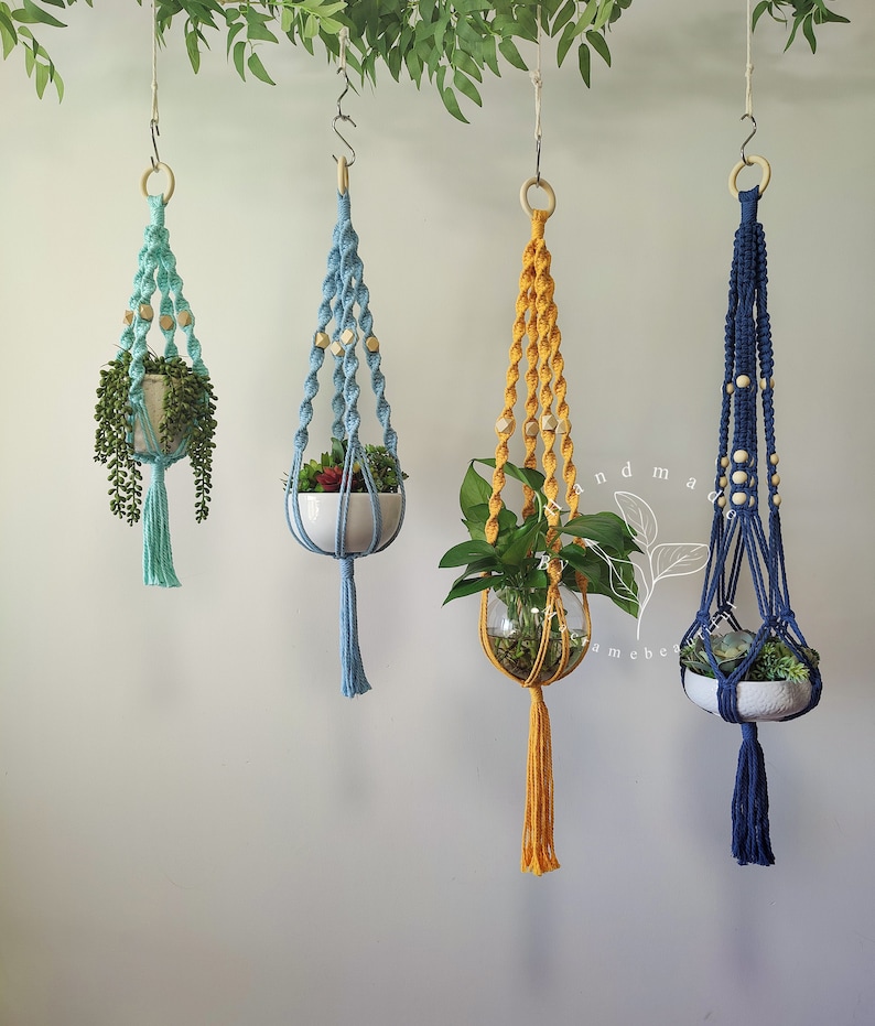 May include: Four macrame plant hangers in shades of teal, blue, yellow, and navy blue, each holding a different plant. The hangers are suspended from wooden rings and feature wooden beads. The plants are in white ceramic pots or glass globes.