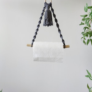 K&ouml;nnte beinhalten: Ein grauer Makramee-Papierhandtuchhalter mit einem Holzstift und einem grauen Quasten, der an einem Holzring h&auml;ngt. Der Halter h&auml;ngt an einem Deckenhaken.
