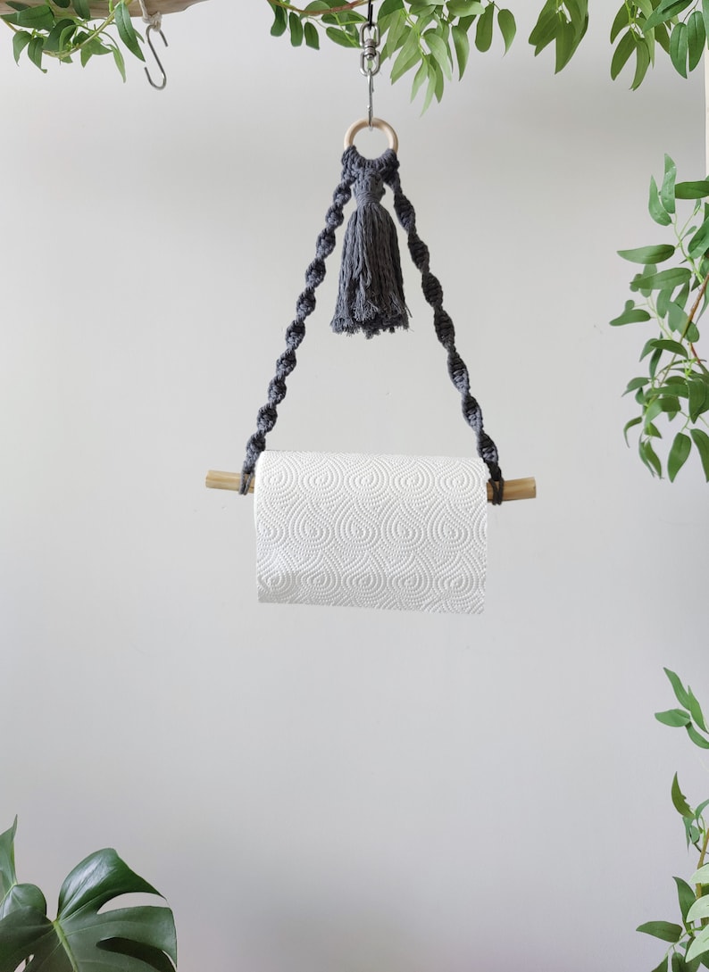 K&ouml;nnte beinhalten: Ein grauer Makramee-Papierhandtuchhalter mit einem Holzstift und einem grauen Quasten, der an einem Holzring h&auml;ngt. Der Halter h&auml;ngt an einem Deckenhaken.