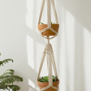 Artisanal macrame plant hanger in style B featuring intricate knotwork and tassels, displaying two terracotta pots with lush greenery, highlighted by natural light and shadows.