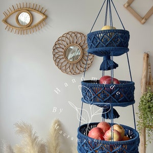 Navy blue 3 tier macrame fruit basket with three circular tiers, ornate weaving, and tassels, hung by a wooden ring, showcasing fresh fruits and plants in a cozy home setting with decorative accents