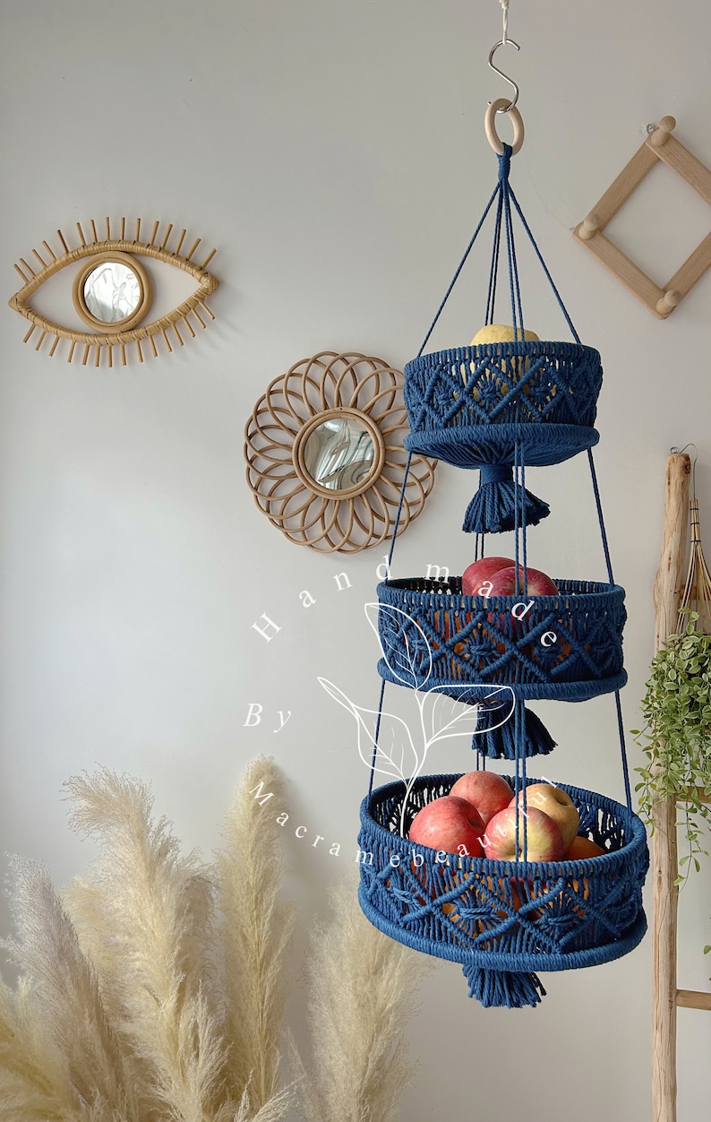 Navy blue 3 tier macrame fruit basket with three circular tiers, ornate weaving, and tassels, hung by a wooden ring, showcasing fresh fruits and plants in a cozy home setting with decorative accents