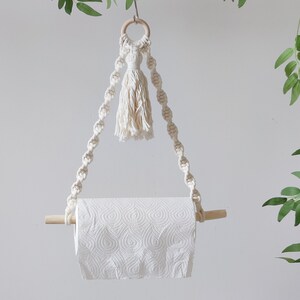 K&ouml;nnte beinhalten: Ein wei&szlig;er Makramee-Papierhandtuchhalter mit einem Holzstift und einem Quasten, der oben h&auml;ngt.