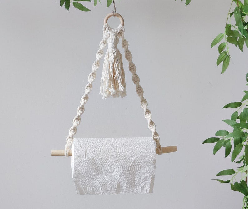 K&ouml;nnte beinhalten: Ein wei&szlig;er Makramee-Papierhandtuchhalter mit einem Holzstift und einem Quasten, der oben h&auml;ngt.