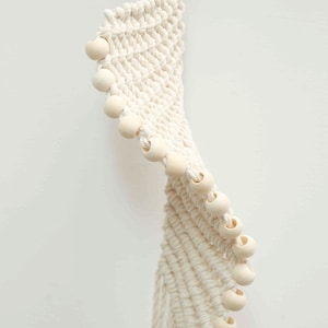May include: Cream-colored macrame plant hanger with wooden beads. The woven design features a spiral pattern, with a wooden ring at the top for hanging. The beads are evenly spaced along the edge of the macrame.