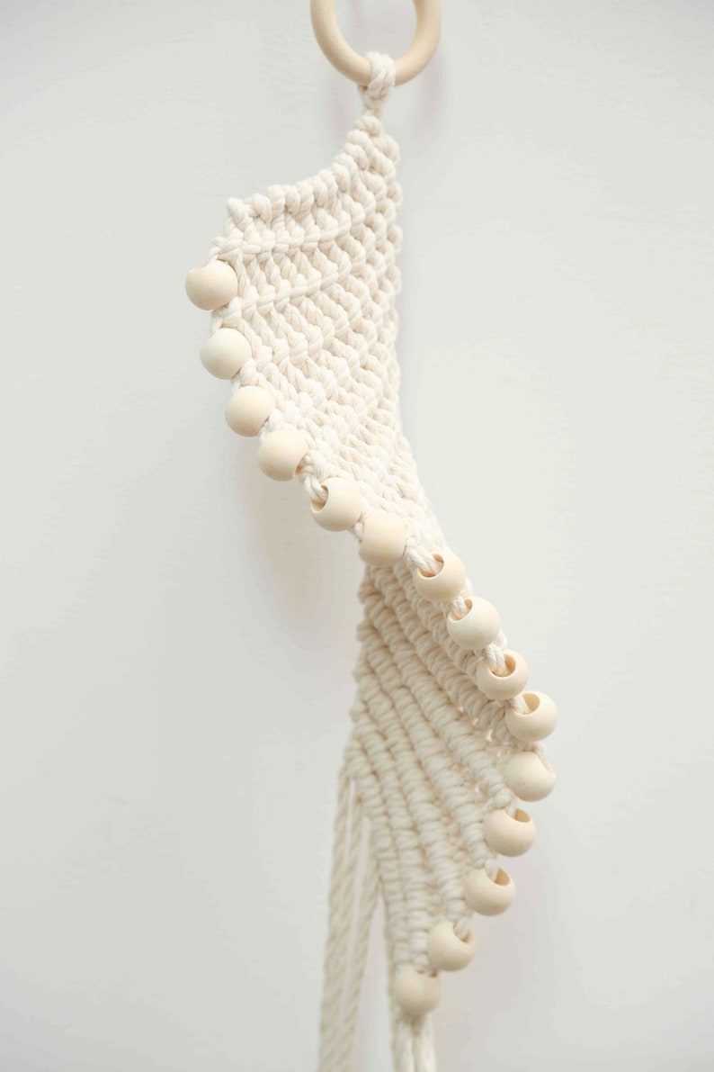 May include: Cream-colored macrame plant hanger with wooden beads. The woven design features a spiral pattern, with a wooden ring at the top for hanging. The beads are evenly spaced along the edge of the macrame.