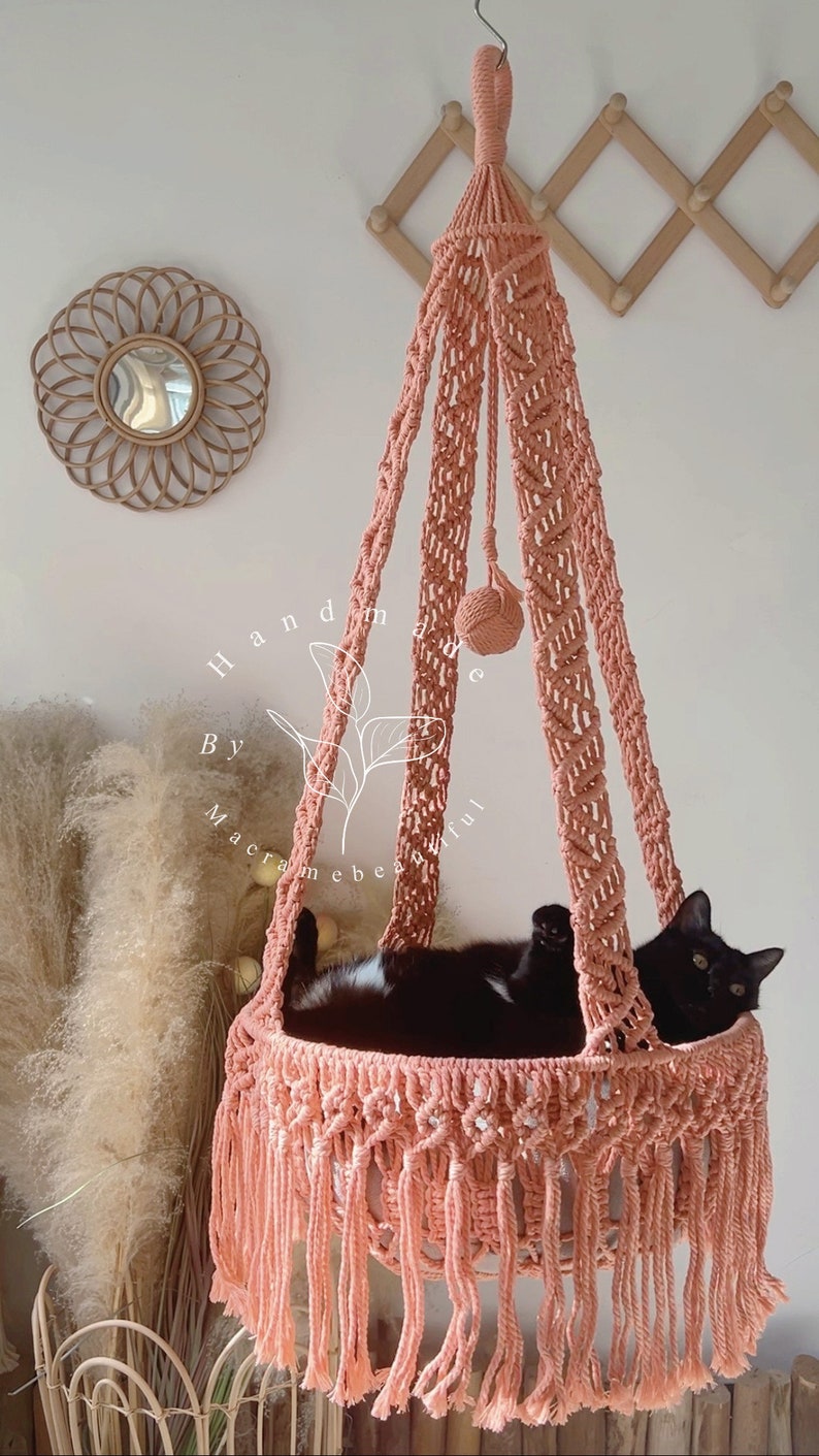 May include: A peach-colored macrame cat bed with a black cat inside. The hanging bed has a woven design and fringe at the bottom. The text "handmade by macramebeautiful" is visible on the side.