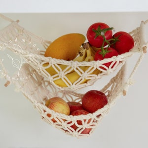 Overhead view of a two-tiered macrame fruit hammock filled with fresh bananas, tomatoes, and apples, showcasing the detailed knotwork, providing a stylish and bohemian kitchen storage solution