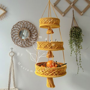 Three-level handmade yellow macrame hanging fruit basket filled with oranges and other produce, against a boho decor background