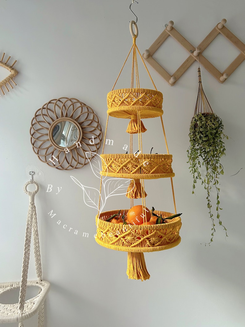 Three-level handmade yellow macrame hanging fruit basket filled with oranges and other produce, against a boho decor background
