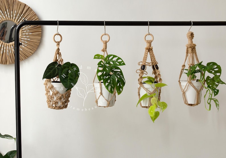 Handmade jute macrame plant hangers with wooden rings, featuring four unique knotted designs suspending white ceramic pots filled with lush green trailing plants—boho indoor décor ideal for hanging planters, balconies, patios and cozy living spaces.