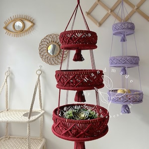 Vibrant red macrame fruit basket with three circular tiers, ornate weaving, and tassels, hung by a wooden ring, showcasing fresh fruits and plants in a cozy home setting with decorative accents