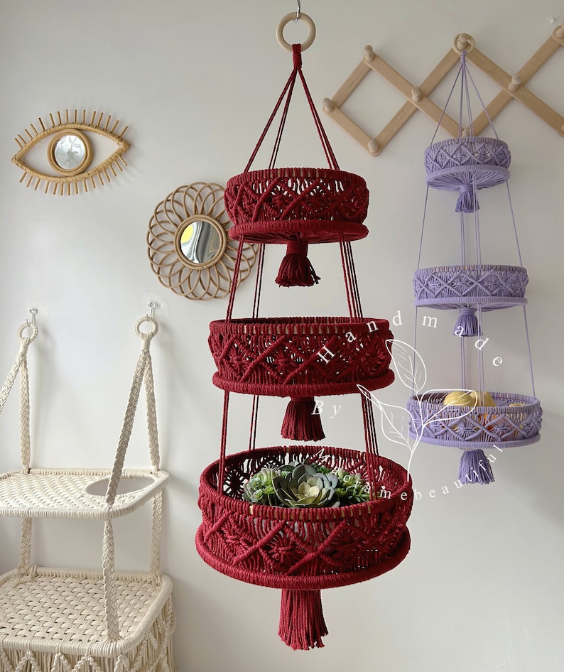 Vibrant red macrame fruit basket with three circular tiers, ornate weaving, and tassels, hung by a wooden ring, showcasing fresh fruits and plants in a cozy home setting with decorative accents