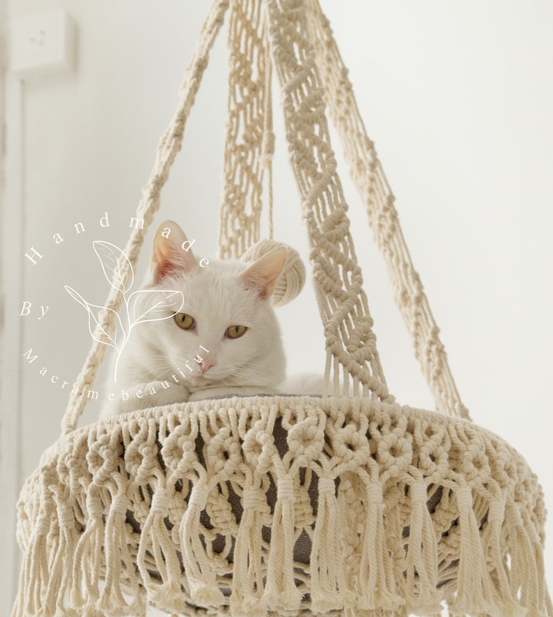 May include: A cream-colored macrame cat bed with a white cat resting inside. The bed is suspended from the ceiling and features intricate knotting and fringe details. The text "Handmade By Macrame Beautiful" is visible on the left side.