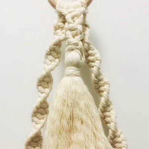 K&ouml;nnte beinhalten: Ein wei&szlig;er Makramee-Pflanzenh&auml;nger mit einem Holzring oben und einem langen Quasten unten.