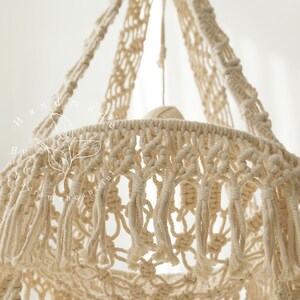 May include: Close-up of a cream-colored macrame hanging basket. The basket is intricately woven with knotted patterns and features long fringe along the bottom edge. The text "Handmade by macrame beautiful" is visible on the left side.