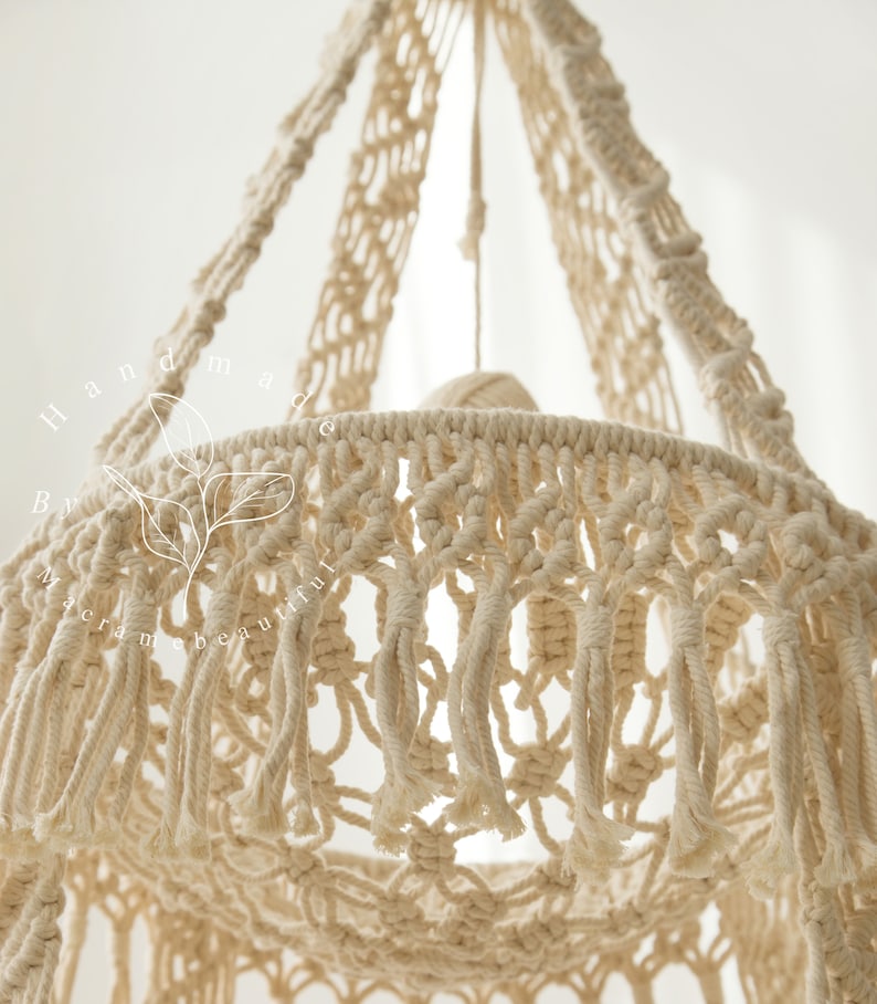 May include: Close-up of a cream-colored macrame hanging basket. The basket is intricately woven with knotted patterns and features long fringe along the bottom edge. The text "Handmade by macrame beautiful" is visible on the left side.