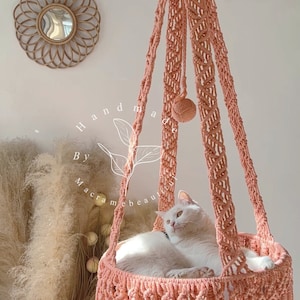 May include: A peach-colored macrame cat bed with a white cat inside. The handmade bed is suspended from the ceiling and features a woven design with fringe. The text "Handmade By macramebeautiful" is visible.