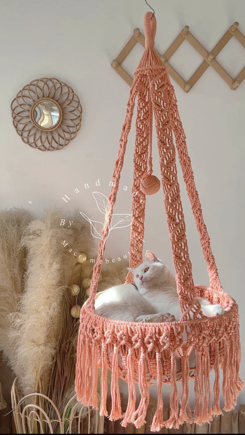 May include: A peach-colored macrame cat bed with a white cat inside. The handmade bed is suspended from the ceiling and features a woven design with fringe. The text "Handmade By macramebeautiful" is visible.