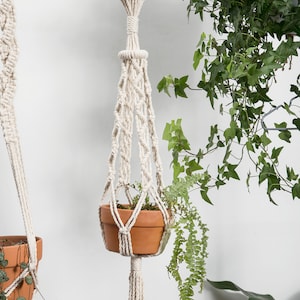 Style D macrame plant hanger, 100 cm long with tassels, handcrafted in a natural white hue, featuring detailed knots and a hanging terracotta pot with trailing greenery, enhancing indoor decor.