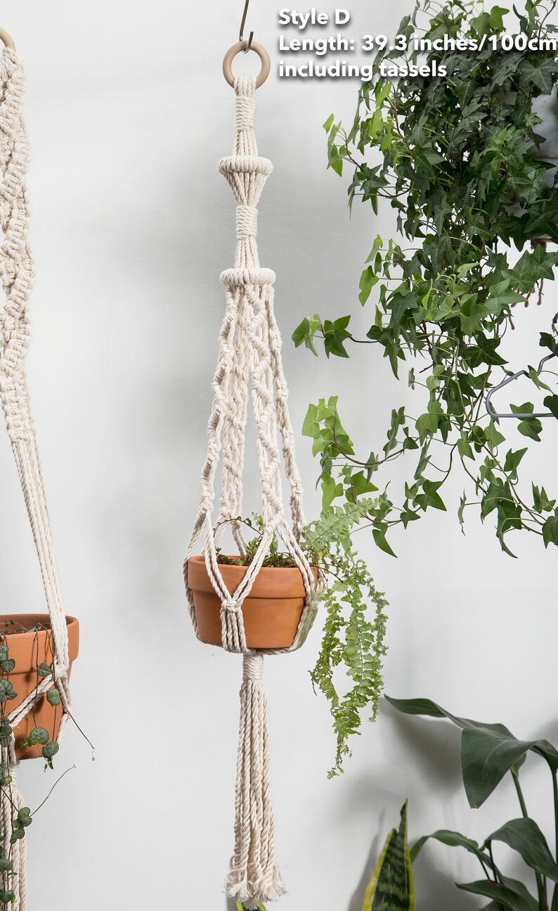 Style D macrame plant hanger, 100 cm long with tassels, handcrafted in a natural white hue, featuring detailed knots and a hanging terracotta pot with trailing greenery, enhancing indoor decor.