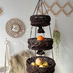 Brown macrame fruit basket with three circular tiers, ornate weaving, and tassels, hung by a wooden ring, showcasing fresh fruits Boho home setting with decorative accents