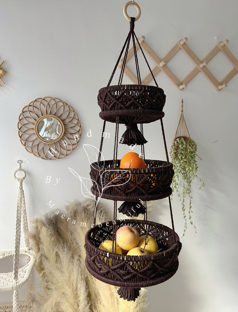 Brown macrame fruit basket with three circular tiers, ornate weaving, and tassels, hung by a wooden ring, showcasing fresh fruits Boho home setting with decorative accents