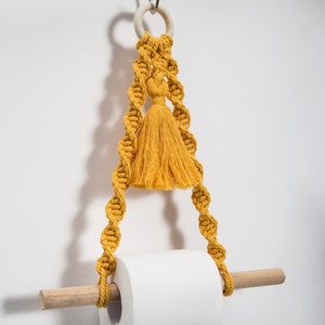 K&ouml;nnte beinhalten: Ein gelber Makramee-Toilettenpapierhalter mit einem Holzstift und einem Quasten, der oben h&auml;ngt.