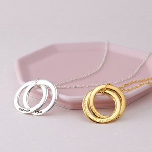 May include: Two personalised necklaces with interlocking circles, one silver and one gold. The silver necklace has the names "Carmen" and "Tom" engraved. The gold necklace has the names "Stanluca" and "Sammy" and the date "12.27.04" engraved.
