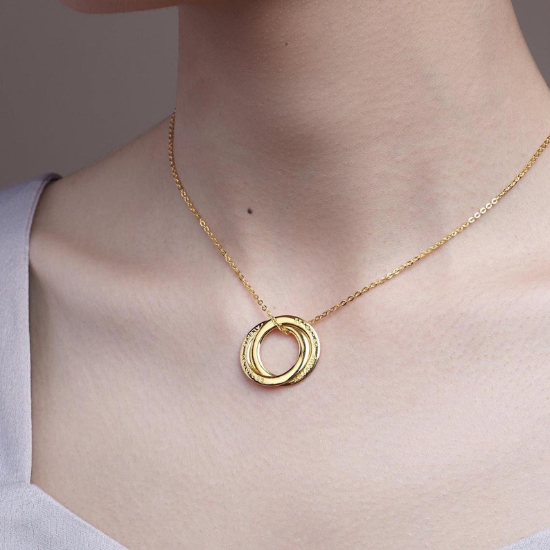 May include: A gold necklace with three interlinked rings as a pendant. The rings are suspended from a fine chain. The necklace is worn around the neck, and the background is a plain colour.