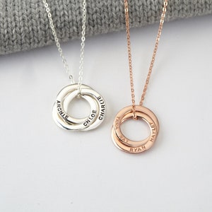 May include: Two personalised necklaces featuring interlocking rings. One is silver, engraved with "ARCHIE", "CHLOE", and "CHARLIE". The other is rose gold, with "TARA", "NAT", "RYAN", and "HAYLEY". Both hang on fine chains.