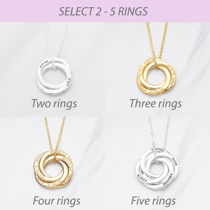 May include: A selection of personalised ring necklaces in silver and gold. The image displays options with two to five rings, each engraved with names and dates. The text at the top reads "SELECT 2-5 RINGS".