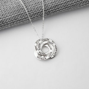 May include: A silver necklace featuring a pendant of interconnected rings, each inscribed with a name. The necklace is shown against a grey knitted background.