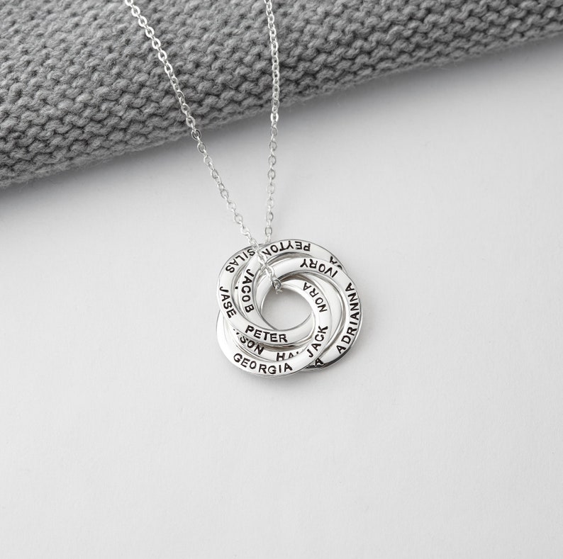 May include: A silver necklace featuring a pendant of interconnected rings, each inscribed with a name. The necklace is shown against a grey knitted background.