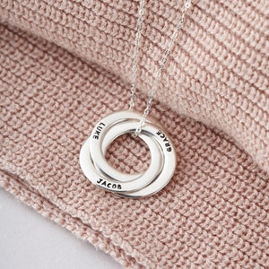 May include: A silver necklace featuring three interlinked rings, each inscribed with a name: Luke, Grace, and Jacob. The necklace is set against a soft, pink knitted backdrop.