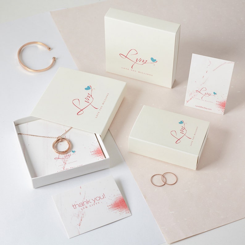 May include: A rose gold jewellery set, including a necklace, bracelet, and rings, presented with gift boxes and cards. The boxes and cards display a logo with the text "Love You Millions". The background is a soft pink and white.