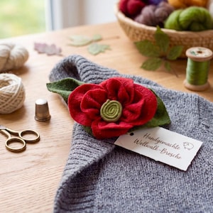 May include: A red felt flower brooch with green felt leaves and a spiral center, attached to a grey knitted garment. A white rectangular tag is attached to the brooch with the text Handgemachte Wollwalk-Brosche and a small sheep icon.
