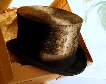 May include: A black top hat with a textured, fur-like finish on the crown and a smooth, solid black band around the base. The hat features a flat top and a curved brim, resting inside an open cardboard box with light brown interior panels.